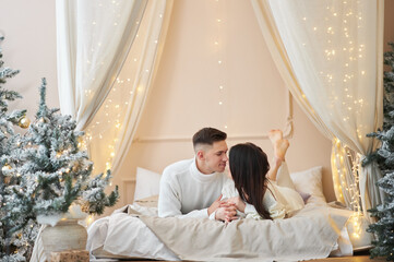 Top view of passionate romantic couple spending time before New Year at home with garlands. Sexy young woman and handsome man are lying on bed in sweaters and hugging. - obrazy, fototapety, plakaty