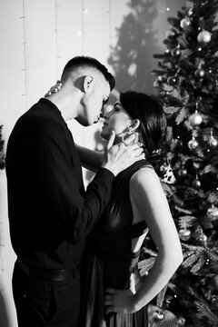 A young couple in love kiss on Christmas Eve by the Christmas tree with gifts at home in December. Black and white photo