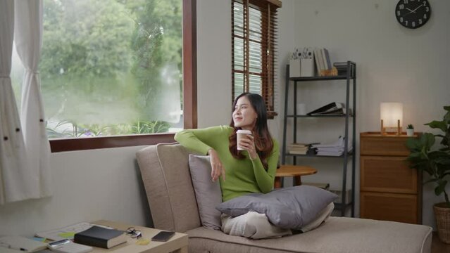 Woman green sweater sitting on a sofa and drinking coffee and looking outside the windows for relaxing and chill out.