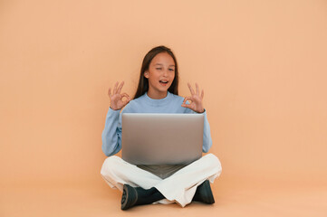 Sitting with laptop. Showing gesture. Cute young girl is in the studio against background