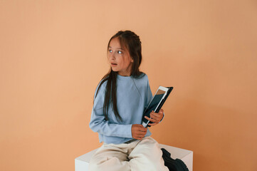 With tablet in hands. Sitting on cube. Cute young girl is in the studio against background