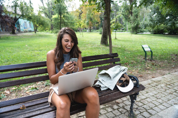 Obraz premium Young freelance modern hipster woman working online in the park sitting on the bench having video call on web cam on computer. E-Commerce independent female small company owner marketing adviser