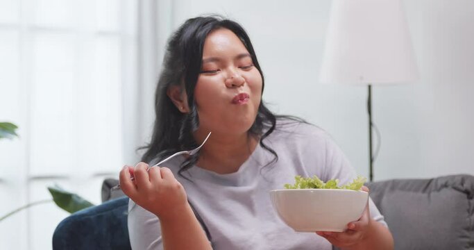 A chubby Asian woman enjoy eating salad  while relaxing on the sofa at home.
