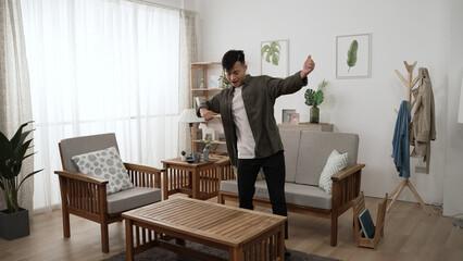 energetic asian young guy in smart casual dancing in a modern home living room interior with...