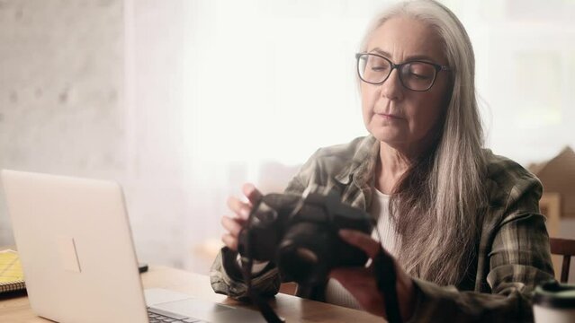 Portrait of beautiful gray haired senior woman photographer hold digital camera looking at screen choosing photos for editing while sitting in front of laptop computer at home workplace