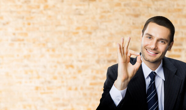 Happy smiling successful gesturing business man with okay hand sign at office brown bricks wall background.