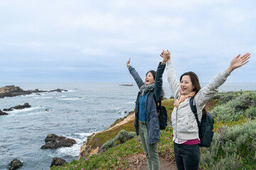 two cheerful asian Taiwanese female travelers cheering and enjoying fresh air and ocean view with raised outstretched arms at shore of big sur California usa - Powered by Adobe