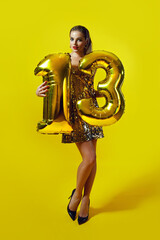 A slender young tanned woman in a shiny dress holds the number 13 folded from golden inflatable balloons. Holiday, birthday. Number 13. Model posing on a golden background.