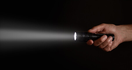 Male hand holding a led flashlight with a wide white beam on a black background, leaving the right...
