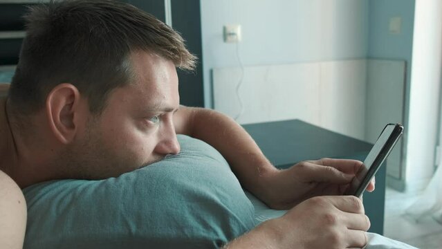 Handsome man using mobile phone in bed at the morning surfing internet, checking social media and reading news. Gadget addiction. Technology concept