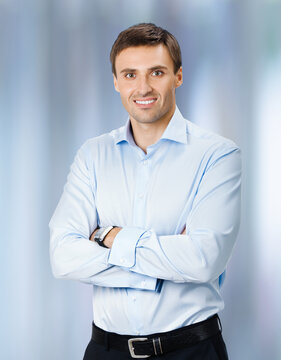 Portrait photo of young executive businessman in confident shirt cloth, crossed arms, on blurred office background. Business concept. Smiling professional office worker, employee man.