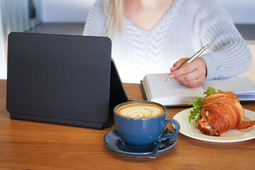 Obraz na płótnie Canvas Close up woman in casual clothes sitting and browsing laptop at kitchen table with cup of coffee and plate of croissant