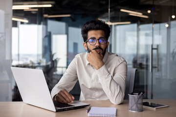 Portrait of a young Indian male student who is studying in the office at the table at the laptop, looking seriously and thoughtfully at the camera - obrazy, fototapety, plakaty