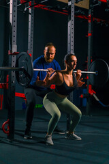 portrait of a young beautiful focused fitness woman doing a heavy shoulder press exercise with a barbell in the gym under the supervision of a trainer