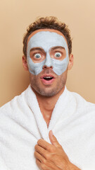 Young man with cream mask and white bath towel on his neck opens eyes widely with surprise standing on studio background, male skincare concept, copy space