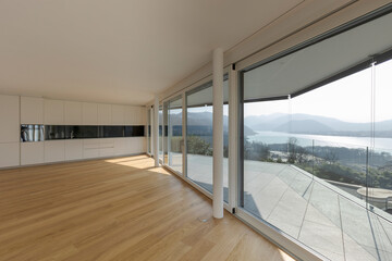 Modern living room with open space and a black and white designed kitchen situated at the end. Large windows allow plenty of sunlight to enter.