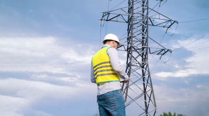 Caucasian male engineer in a helmet and safety glasses uses a smartphone for field work near a telecommunications tower that controls cellular electrical installations.