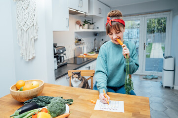 Young woman filling veganuary calendar while eating fresh carrot sitting on kitchen. Fresh...