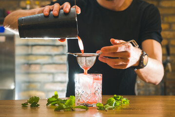Barman gently holds a mixing cup and strainer and pours colorful pink cocktail into a glass on the...