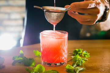 Barman gently holds a mixing strainer and pours colorful cocktail into a glass surrounded by scattered mint leaves on the wooden bar counter