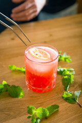 Bartender holding tweezers putting lemon slice over glass with cold cocktail on bar counter with scattered mint leaves on wooden counter
