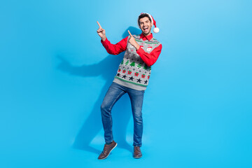 Full body photo of cute young man jump point empty space promo dressed x-mas print vest hat clothes isolated on blue color background