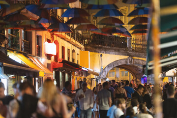 Fototapeta na wymiar Colorful umbrellas over the street of Portugal - people walk around the city in the evening near a cafe