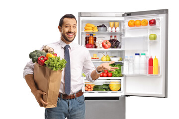 Cheerful young professional man holding a grocery bag in front of an open fridge