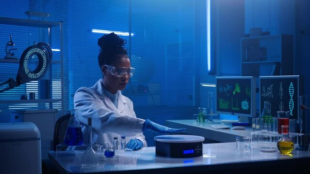 Medium-sized video capturing a dark-skinned female scientist wearing glasses and a lab coat sitting in a laboratory, researching brain hologram displayed in front of her.