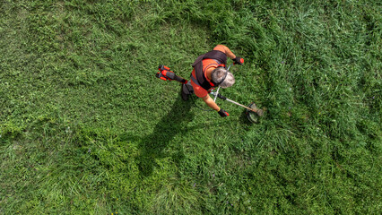 Obraz premium Aerial high angle view of worker in protective clothing cutting grass with brush cutter