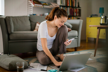 Young woman using her smartphone while going over financials at home