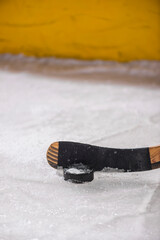 A Ice Hockey puck with a wooden hockey stick with black tape on the blade with the marred up boards and yellow kicker in the background.