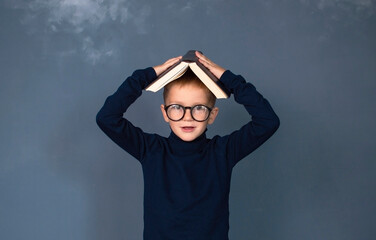 Boy covering his head with book on blue studio background. Portrait of funny smart schoolboy looks...