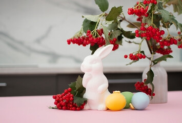 bouquet of mountain ash twigs in vase in home interior and white Easter bunnies on white table. In...