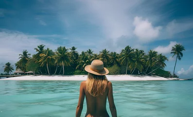 Fototapeten Girl in a hat in the blue water of the ocean in front of an island with palm trees and white sand. © JulMay