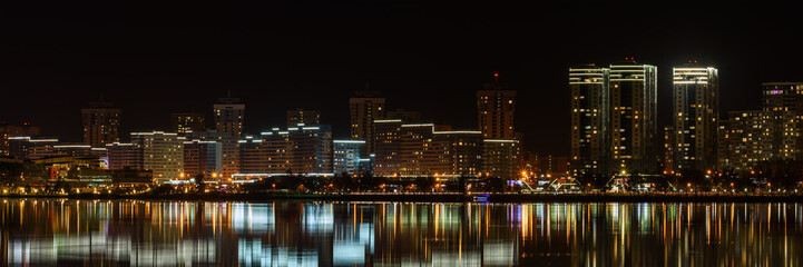 Fototapeta na wymiar modern night big city with tall buildings glows beautifully with electric lights and is reflected on the water of the lake. Coastal Avenue. widescreen panoramic cityscape in 15x3 format. Minsk