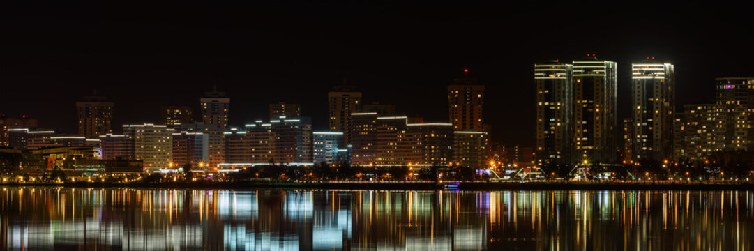 modern night big city with tall buildings glows beautifully with electric lights and is reflected on the water of the lake. Coastal Avenue. widescreen panoramic cityscape in 15x3 format. Minsk