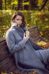 brunette woman in a gray coat and boots is sitting on a park bench in autumn among the trees. Orange foliage and a wooden bench during the day