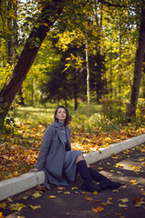 brunette woman in a gray coat and boots is sitting on a park curb in autumn among the trees. Orange foliage and a wooden bench during the day