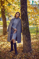brunette woman in a gray coat and boots is standing by a tree in autumn among the trees. Orange foliage during the day