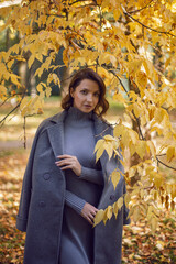 brunette woman in a gray coat and boots is standing by a tree in autumn among the trees. Orange foliage during the day