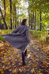 brunette woman in a gray coat and boots is walking along the path in the leaves in autumn among the trees. Orange foliage during the day