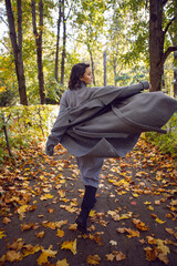 brunette woman in a gray coat and boots is walking along the path in the leaves in autumn among the trees. Orange foliage during the day