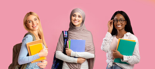 Three multiethnic female students carrying backpacks and workbooks posing over pink background