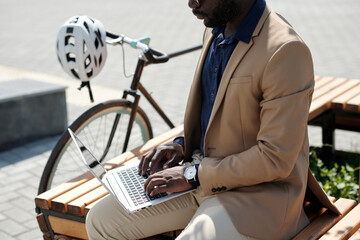 Fototapeta premium Cropped shot of young black man in formalwear typing on laptop keyboard while sitting on wooden bench in front of camera outdoors