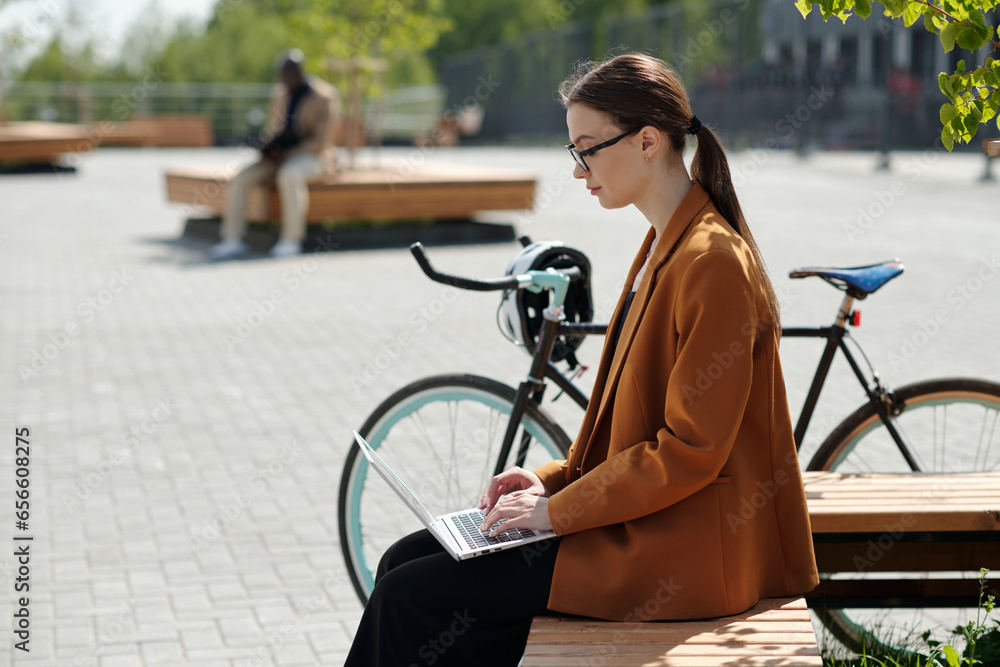 Poster Side view of young serious businesswoman with laptop organizing work while sitting on bench in front of camera and looking at screen