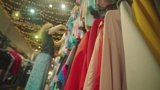 Caucasian woman chooses clothes hanging on a hangers in the cozy vintage second hand shop. Multicolored various second-hand clothes.
