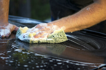 a hand with a washcloth and soap suds washes the hood of the car. Man washing car hood with...