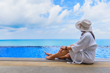 Woman in white bathrobe relaxing at the pool looking at sea view. - obrazy, fototapety, plakaty