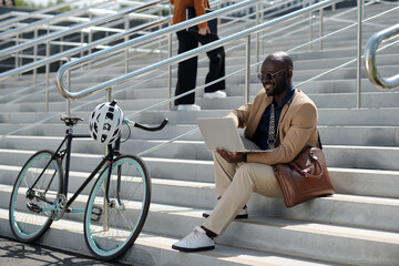 Young successful entrepreneur in formalwear looking at laptop screen while sitting on staircase in urban environment and networking